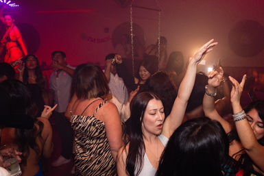 Crowded nightclub dance floor with partygoers dancing under red and purple lights, woman in the foreground singing with her arm raised.