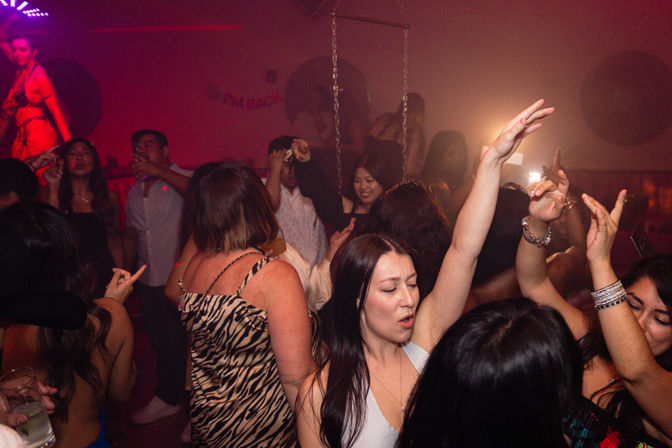 Crowded nightclub dance floor with partygoers dancing under red and purple lights, woman in the foreground singing with her arm raised.