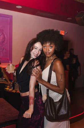 Two friends posing in a pink-lit nightclub lounge — cocktail in hand, evening outfits, visible tattoos and handbag, relaxed party vibe