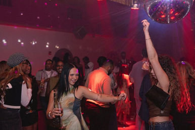 Lively nightclub dance floor with people dancing under a mirrored disco ball, red lighting and guests holding drinks.
