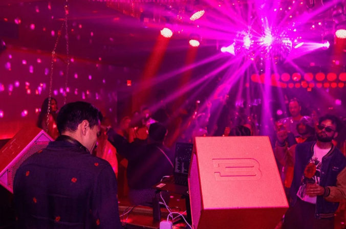 DJ at a booth overlooking a packed nightclub dance floor bathed in pink-purple lights and disco ball reflections, crowd dancing and holding drinks