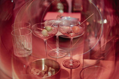 Pink-lit bar table with two martini glasses garnished with olives and a cherry, an empty rocks glass and a bowl of mixed nuts in soft bokeh