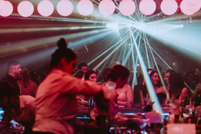 Urban nightclub bar scene at night with bartenders serving drinks, patrons chatting at the bar, a disco ball and laser beams lighting up the dance floor