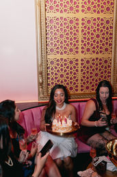 Birthday celebration in a pink velvet booth: woman holding lit cake with candles while friends toast with champagne beneath a decorative gold lattice wall panel.