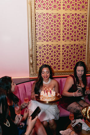 Birthday celebration in a pink velvet booth: woman holding lit cake with candles while friends toast with champagne beneath a decorative gold lattice wall panel.
