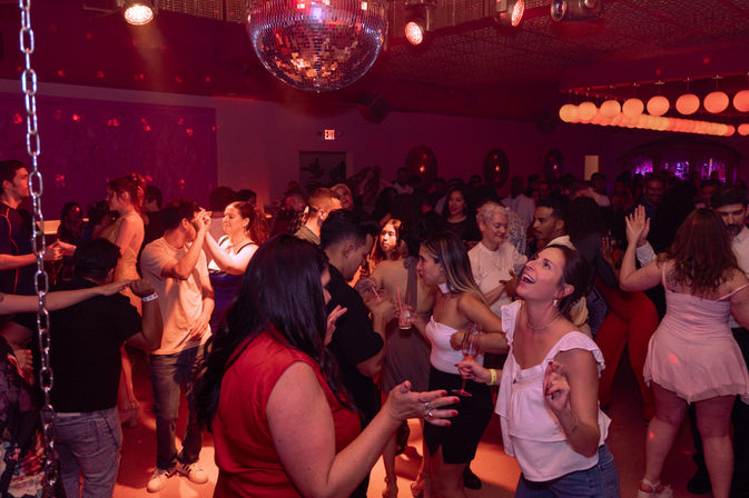 Crowded nightclub dance floor at a late-night party, people dancing and holding drinks under a mirrored disco ball and warm red lighting.