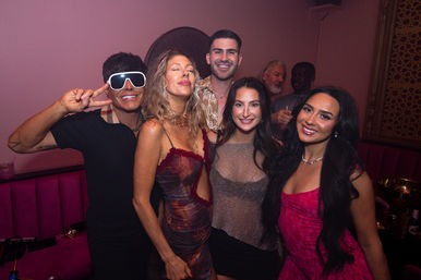 Small group of friends smiling and posing for a photo at a pink-lit nightclub, stylish cocktail dresses and playful oversized sunglasses for a lively night out.