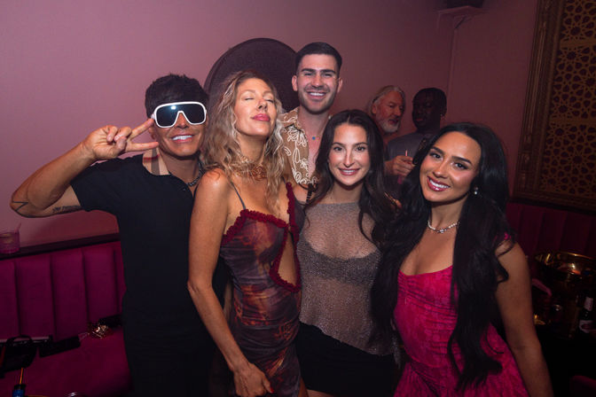 Small group of friends smiling and posing for a photo at a pink-lit nightclub, stylish cocktail dresses and playful oversized sunglasses for a lively night out.
