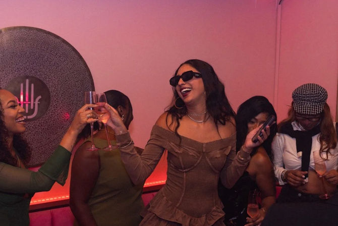 Friends toasting with champagne in a pink-lit lounge — laughing woman in an off-shoulder brown dress and sunglasses holds a phone while others cheer.