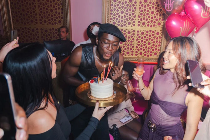 Friends at a trendy pink lounge celebrating a birthday: person blowing out candles on a small frosted cake on a gold tray while others toast with cocktails and take photos.