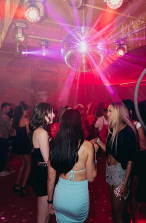 Group of friends socializing on a crowded nightclub dance floor beneath a sparkling disco ball with pink and purple club lights, vibrant city nightlife party scene.