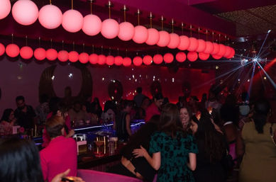 Vibrant nightclub interior at night with rows of glowing pink globe pendant lights, a disco ball scattering beams, a crowded bar and groups of people chatting, dancing and holding drinks.