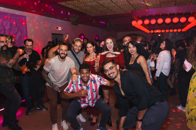 Group of smiling young adults posing on a crowded nightclub dance floor under red lantern lights, holding drinks and dancing — upbeat urban nightlife party scene.