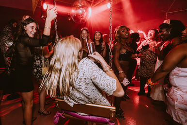 Friends partying under a disco ball in a red-lit nightclub — woman on a swing sipping a cocktail as a diverse crowd dances and cheers.