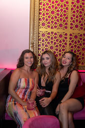 Three women enjoying cocktails in a pink-lit lounge booth, wearing colorful dresses with a decorative gold patterned wall behind them.
