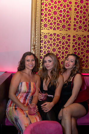 Three women enjoying cocktails in a pink-lit lounge booth, wearing colorful dresses with a decorative gold patterned wall behind them.