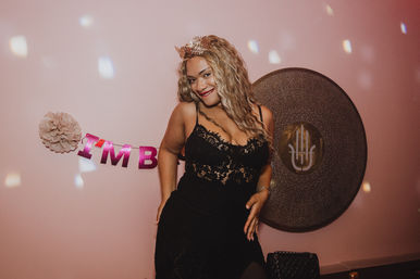 Smiling woman in a black lace dress and tiara posing at an indoor pink party backdrop with a round metallic wall decoration, pink letter banner, and scattered confetti lights.