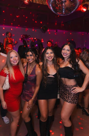 Four smiling women in stylish club outfits posing under a disco ball on a crowded urban nightclub dance floor with red light dots.