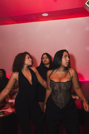 Three women in black outfits dancing and posing in a pink neon-lit lounge, one wearing a lace corset — energetic nighttime party scene.