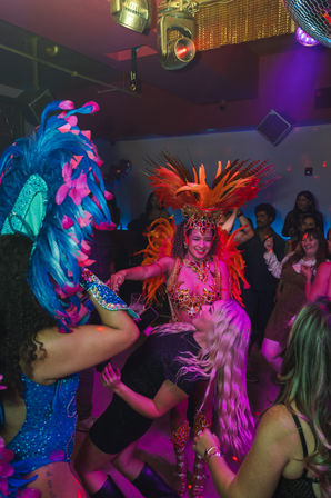 Vibrant nightclub party with carnival-style dancers in colorful feathered headdresses and a crowd dancing under a disco ball