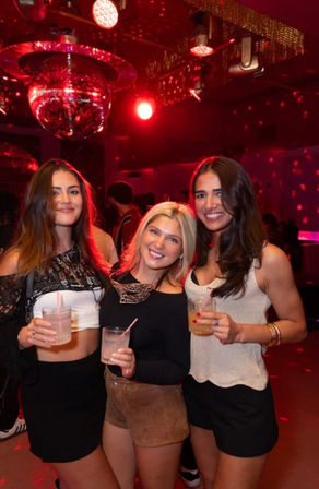 Three friends smiling on a nightclub dance floor under a mirrored disco ball and red lighting, holding cocktails — lively city nightlife scene.