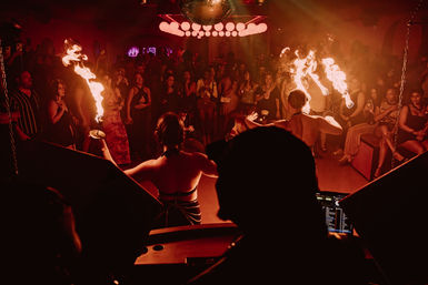 DJ-view inside a packed nightclub: two fire performers twirling flaming props under a disco ball as a cheering crowd watches in moody red lighting.