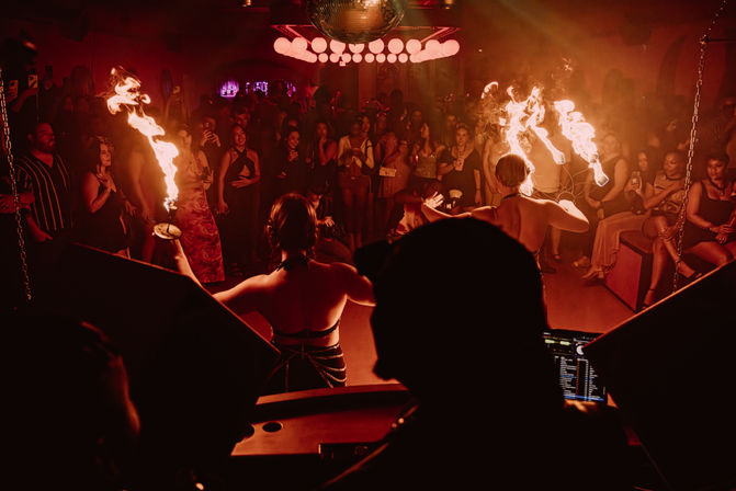 DJ-view inside a packed nightclub: two fire performers twirling flaming props under a disco ball as a cheering crowd watches in moody red lighting.