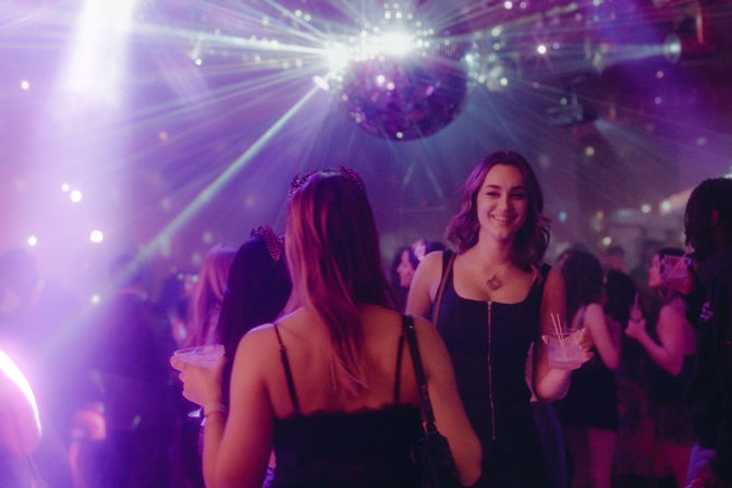 Smiling woman in a black dress holding a cocktail on a crowded dance floor under a glittering disco ball and purple club lights