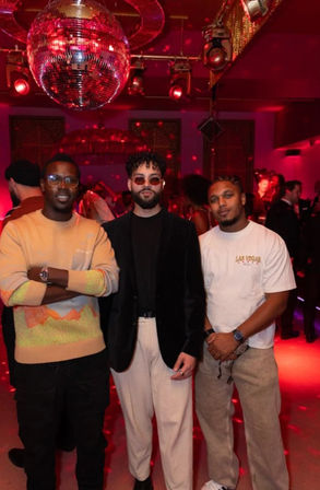 Three men posing under a mirrored disco ball in a red-lit nightclub, wearing stylish casual outfits—one in a Las Vegas tee—with a dancing crowd in the background.