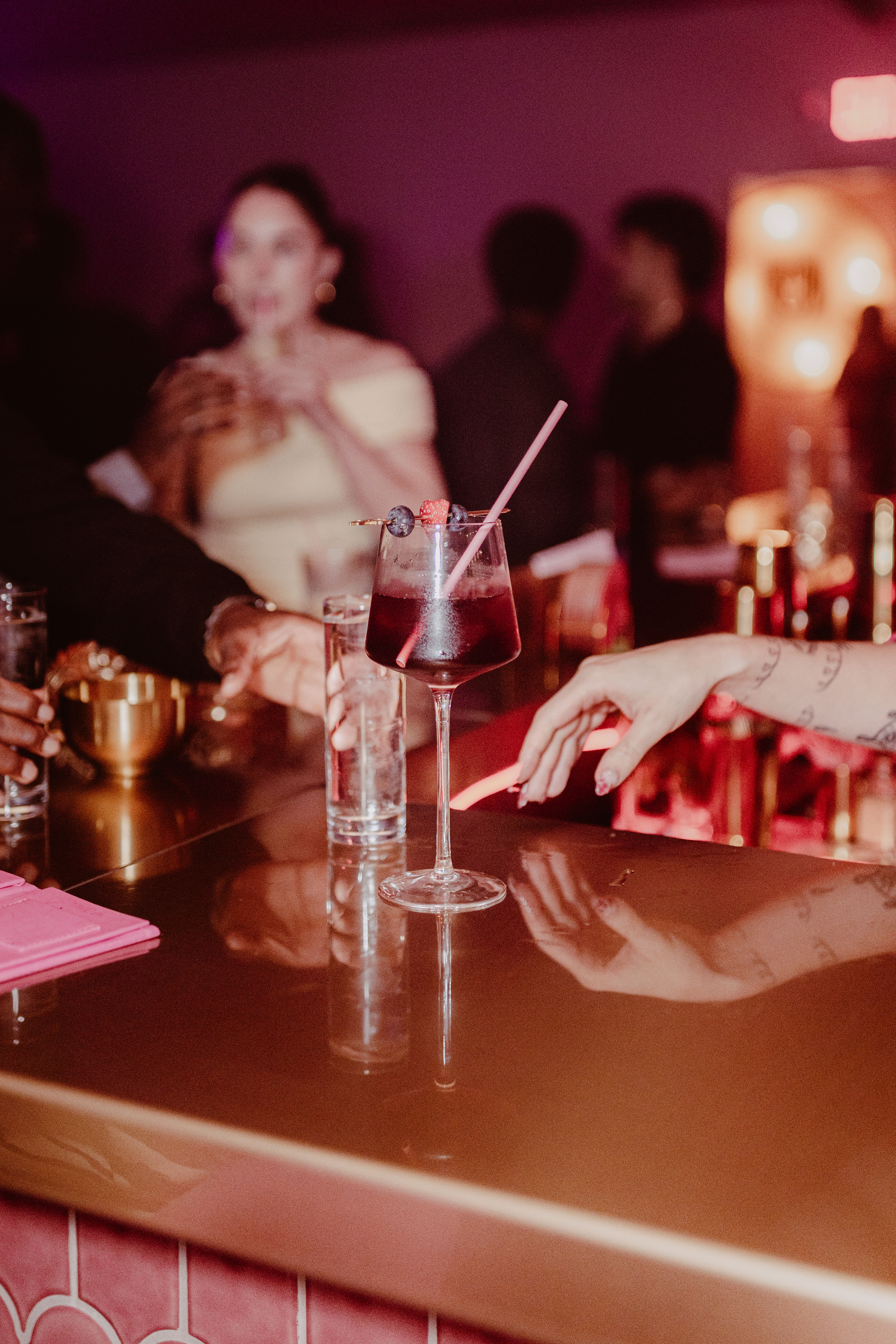 Deep-red berry cocktail in a stemmed glass with a pink straw and berry garnish on a glossy bar counter, hands reaching, pink neon-lit trendy bar nightlife scene.