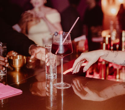 Deep-red berry cocktail in a stemmed glass with a pink straw and berry garnish on a glossy bar counter, hands reaching, pink neon-lit trendy bar nightlife scene.