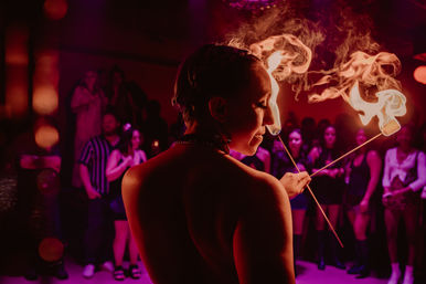 Fire performer in a dim purple-red nightclub, seen from behind, twirling two flaming wands with smoky trails while a crowd watches.