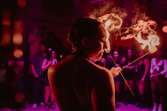 Fire performer in a dim purple-red nightclub, seen from behind, twirling two flaming wands with smoky trails while a crowd watches.