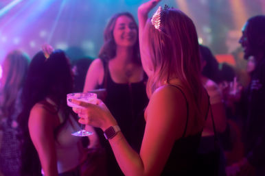 Woman holding a coupe glass with a cocktail, sparkly hair clip and smartwatch glinting under pink-and-blue nightclub lights while dancing with friends on a crowded dance floor.