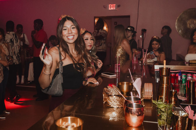 Smiling women wearing tiaras at a crowded indoor bar counter with cocktails, gold accents and lively nightlife party vibe.