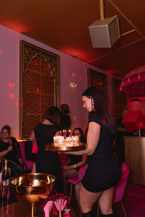 Woman carrying lit birthday cake through an upscale pink-and-gold lounge with velvet seating and partygoers, festive nightlife vibe