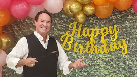 Smiling man in white shirt and black vest posing at a birthday party backdrop with a yellow "Happy Birthday" sign and pink, gold and orange balloons over a leafy wall.