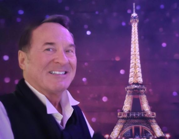 Smiling man posing beside an illuminated Eiffel Tower backdrop with purple bokeh lights in a playful Paris-themed photo.