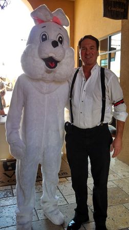 Smiling man in a white dress shirt and black suspenders posing beside a fluffy white Easter bunny costume with a pink bow in a sunlit hotel lobby entrance on tiled floor.