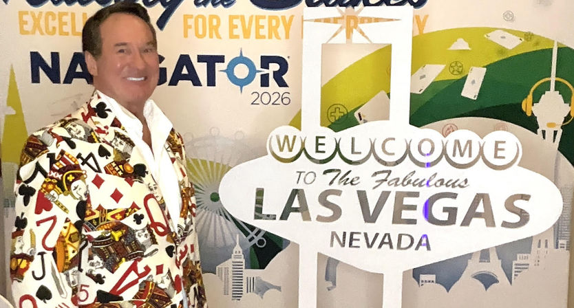 Smiling event attendee in a playing-card patterned blazer poses beside a Welcome to the Fabulous Las Vegas Nevada sign cutout against a conference backdrop with 2026 branding.