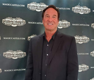 Smiling man in a black suit posing in front of a vintage magic-themed step-and-repeat event backdrop