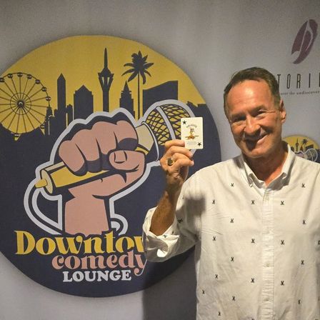 Smiling man holding a Joker card in front of a colorful comedy club backdrop featuring a fist gripping a microphone, city skyline and Ferris wheel.
