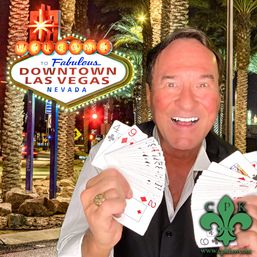 Smiling magician holding fanned playing cards in front of the illuminated "Welcome to Fabulous Downtown Las Vegas" sign at night with palm trees and city lights