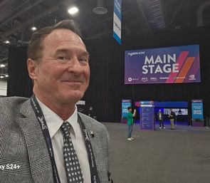Man smiling in a plaid suit takes a selfie inside a large conference expo hall with a colorful "MAIN STAGE" banner and a few attendees in the background.