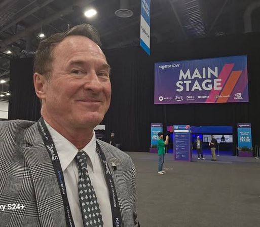 Man smiling in a plaid suit takes a selfie inside a large conference expo hall with a colorful "MAIN STAGE" banner and a few attendees in the background.