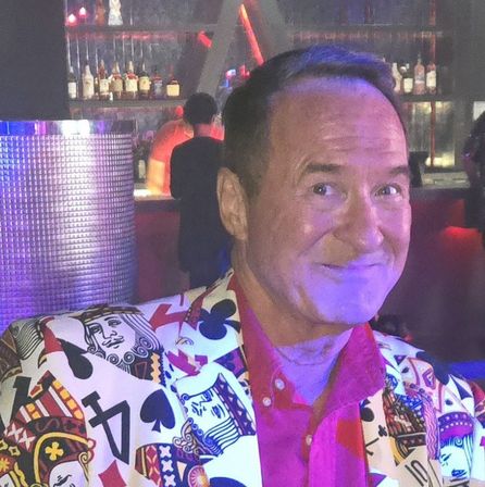 Smiling man wearing a vibrant playing-card patterned blazer and pink shirt under purple neon lights at a nightclub bar with liquor bottles on shelves.