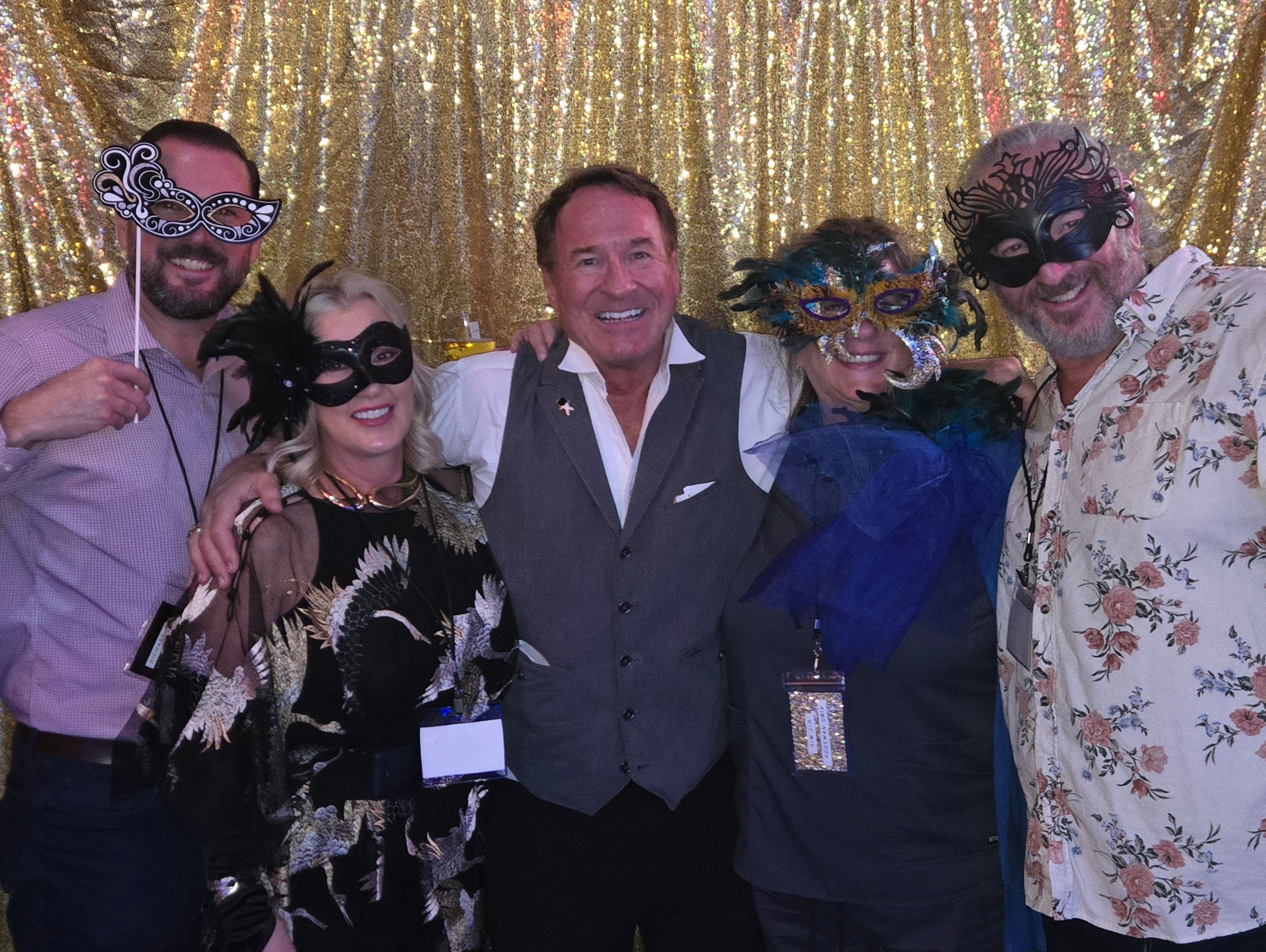 Five adults in festive attire wearing colorful masquerade masks and holding props, smiling together in a group photo in front of a gold sequin backdrop at an indoor party.