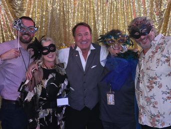 Five adults in festive attire wearing colorful masquerade masks and holding props, smiling together in a group photo in front of a gold sequin backdrop at an indoor party.