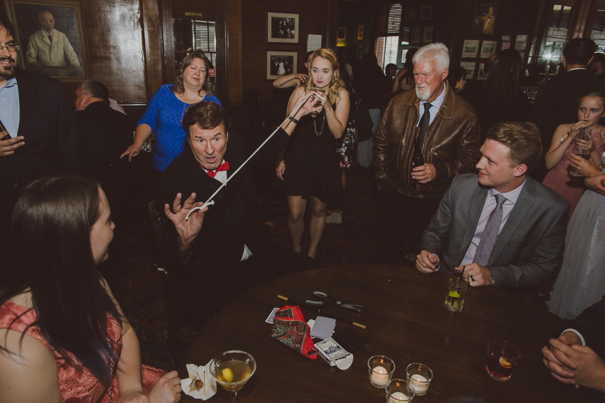 Table-side magician in a wood-paneled lounge entertains party guests with a card trick while onlookers hold drinks around a candlelit table.