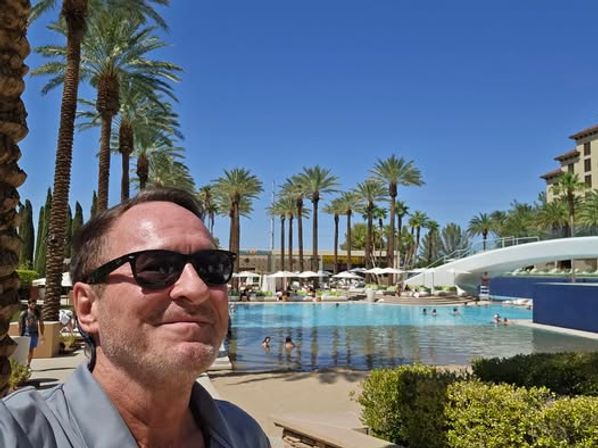 Selfie of a man wearing sunglasses at a sunny palm-tree–lined resort pool with lounge chairs, a white pool bridge and clear blue sky.